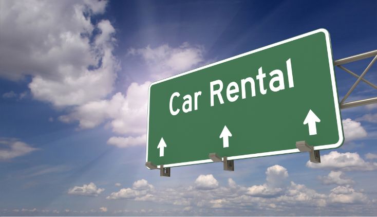 A green road sign that says "car rental" against a blue sky with white clouds.