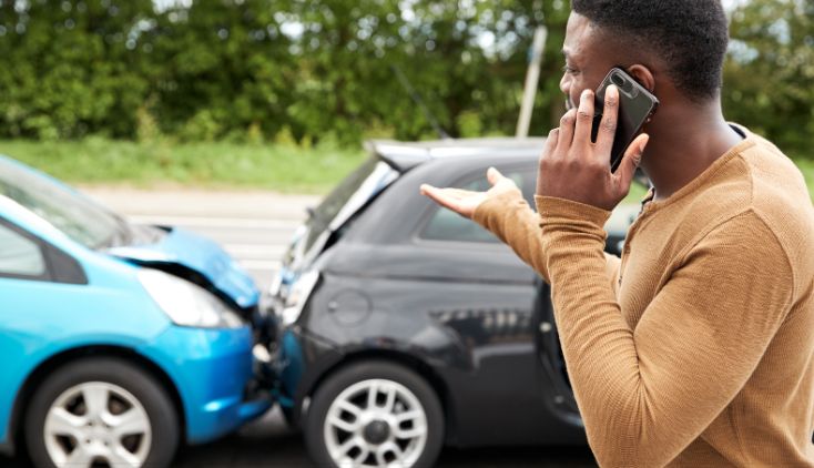 A blue and black car in a fender bender accident. A man is standing in front of the cars on his cell phone.